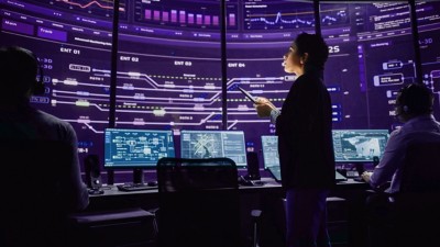 Young Multiethnic Female Government Employee Uses Tablet Computer in System Control Monitoring Center. In the Background Her Coworkers at Their Workspaces with Many Displays Showing Technical Data.