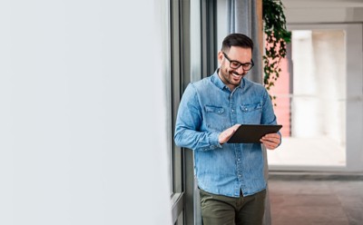 Smiling man businessman in casuals standing in office next to window using tablet making online shopping purchasing Small business home office entrepreneur looking at tablet making online video call