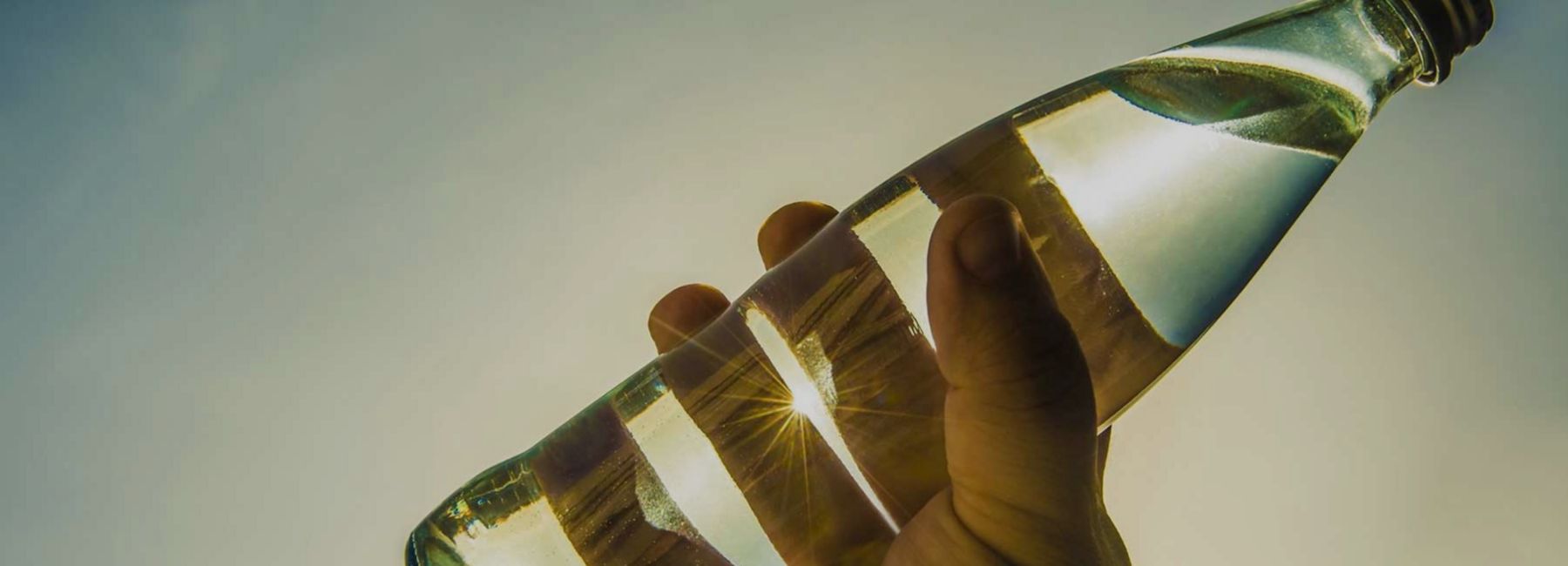 Hand holding glass bottle against blue sky and bright sun, software solutions for manufacturing industry, O-I Glass | DXC Technology Customer Stories